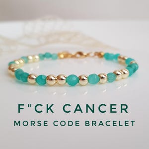 May include: A delicate bracelet featuring alternating gold-colored beads and turquoise-colored beads. The bracelet is inscribed with the text "F"CK CANCER MORSE CODE BRACELET" in dark green letters.