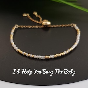 May include: Gold-toned adjustable bracelet with a mix of gold and clear beads. The bracelet has a sliding knot closure and a delicate chain. The text "I'd Help You Bury The Body" is written in white script below the bracelet.