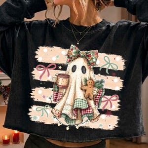 May include: A black long-sleeve sweatshirt featuring a graphic of a ghost wearing a large floral bow, holding a mug and a gingerbread cookie. The ghost is draped in a patchwork-style fabric with red, green, and white patterns over a light background.