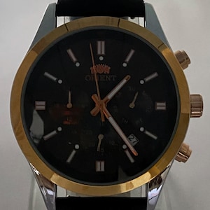 May include: An Orient wristwatch with a black dial, gold-tone bezel, and silver-tone case. The watch features rose gold-tone hands, hour markers, and a black leather strap. A date window displays the number 27, and the Orient logo is visible.