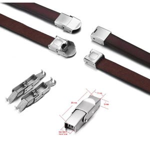 Pode incluir: Close-up de pulseiras de relógio e fechos. Pulseiras de couro castanho com fechos de metal prateado. Os fechos são retangulares com um design articulado. Dimensões do fecho: 25 mm de comprimento, 7 mm de largura e 5 mm de espessura.