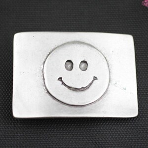 Op de afbeelding: Zilverkleurige rechthoekige gesp met een verhoogd smiley-gezicht ontwerp. Het smiley-gezicht heeft twee kleine oogindrukken en een gebogen mond. De gesp is gemaakt van glanzend metaal.