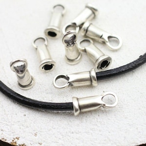 Può includere: Terminali per cordoncino in metallo argentato con un anello per la creazione di gioielli. Diversi terminali sono sparsi, alcuni attaccati a un cordoncino nero. Questi terminali vengono utilizzati per rifinire le estremità di cordoncini o pelle.