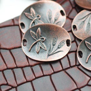 May include: Oval-shaped, antique copper-colored metal embellishments. Each features a detailed dragonfly and wheat stalk design. The embellishments have small holes on each end for attachment. The background is a textured, dark brown surface.