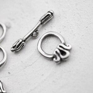 May include: Close-up of a silver-colored toggle clasp jewelry finding. The clasp features a bar and a decorative ring with ornate detailing. The metal has an antique finish, suggesting a vintage or handcrafted style. Ideal for jewelry making.