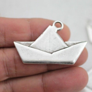 Puede incluir: Un dije de barco de papel plateado con un lazo en la parte superior para sujetarlo a un collar o pulsera. El dije es una réplica en miniatura de un barco de papel plegado, con pliegues y dobleces detallados. Ideal para la creación de joyas.