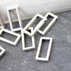 Puede incluir: Una colección de pequeños dijes rectangulares de metal plateado. Los dijes tienen un marco abierto, con un diseño geométrico simple. Están dispersos sobre una superficie gris oscuro, creando un contraste sutil. Estos dijes son adecuados para la fabricación de joyas.