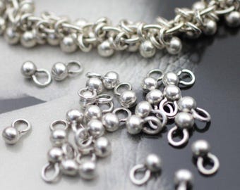 Könnte beinhalten: Nahaufnahme von silberfarbenen Schmuckzubehörteilen, darunter kleine Kugelperlen mit Schlaufen und ein kettenartiges Armband. Die Zubehörteile sind auf einer reflektierenden Oberfläche verstreut, was ihren metallischen Glanz zeigt.