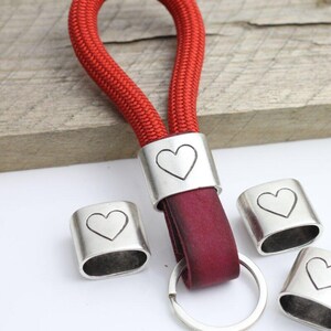 May include: A red rope keychain with a burgundy leather strap and a silver-colored heart-shaped accent. Several silver-colored heart-shaped accents are scattered around the keychain. The keychain has a silver-colored key ring.