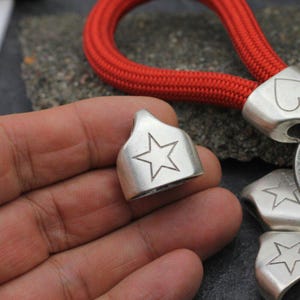 May include: Silver-colored metal tag with a star design. The tag is held in a hand, with a red rope in the background. Other tags with star and heart designs are also visible.