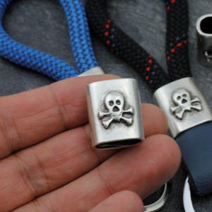 May include: A silver-colored metal clasp with a skull and crossbones design. The clasp is rectangular and appears to be attached to a blue and black rope. The skull and crossbones are raised and detailed.