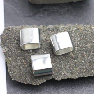 May include: Three shiny, silver-colored rectangular metal beads. Each bead has a smooth, reflective surface and a rounded, hollow interior. The beads are displayed on a rough, gray stone surface.