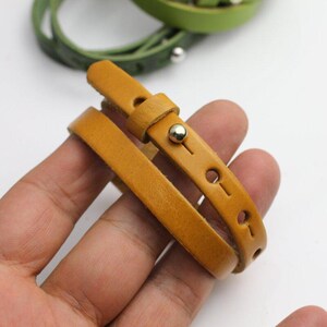 May include: A mustard yellow leather wrap bracelet featuring a smooth finish, a silver-toned metal stud closure, and multiple adjustment holes along the strap. A second green leather bracelet is visible in the blurred background.