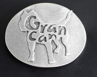 May include: A circular, silver-toned metal emblem. It features a dog silhouette with the words "Gran Can" cut out. The emblem has a slightly aged look and is set against a dark background, suggesting a vintage or antique style.