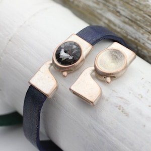 May include: A navy blue leather bracelet with a silver-tone clasp. The clasp features a large, dark gray crystal and a matching empty setting. The bracelet is shown unclasped, displaying the details of the closure mechanism.