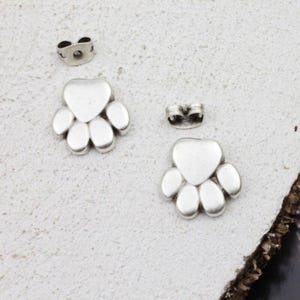 Op de afbeelding: Zilverkleurige oorringen met pootafdrukken en bijpassende oorbelruggen. De oorbellen hebben de vorm van een poot met een hartvormige bovenkant en vijf afgeronde kussentjes. Op een getextureerd, lichtgekleurd oppervlak.