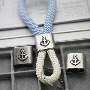 May include: A nautical-themed keychain with a blue and white rope loop and silver-colored metal accents. The metal pieces feature an anchor design. The keychain includes a keyring and two additional anchor-themed metal pieces.