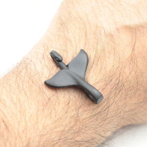 Könnte beinhalten: Ein dunkelgrauer Anhänger in Form einer Walflosse. Der Anhänger besteht aus einem glatten Material und hat oben eine kleine Schlaufe für eine Kette oder ein Band. Das Design ist schlicht und elegant.