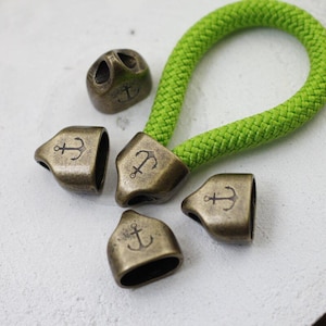May include: Antique bronze-colored metal cord ends and a single bead with an anchor design. A bright green rope is attached to one of the cord ends. Nautical-themed jewelry findings.