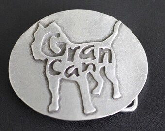 May include: A silver-coloured belt buckle featuring the words "Gran Can" cut out in the shape of a dog. The buckle is round and has a standard clasp.