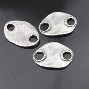 May include: Three silver-toned, oval-shaped metal components with two circular holes each. The components have a slightly textured surface and are arranged on a dark background. The design is simple and modern.