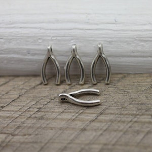 Peut inclure: Quatre breloques en forme de vœux argentés. Trois sont dressées et une est horizontale. Les breloques sont en métal brillant et sont posées sur une surface en bois et un fond en bois peint en blanc.