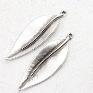 May include: Two silver-toned leaf charms with a distressed finish. Each charm has a central vein detail and a small loop at the top for attaching to jewelry. The leaves are elongated and have a slightly curved shape.
