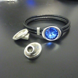 May include: A black leather bracelet with a silver clasp and a large, oval, blue gemstone. Two silver ring-shaped components and a silver oval component are also present, all on a dark surface. The bracelet is a jewelry item.