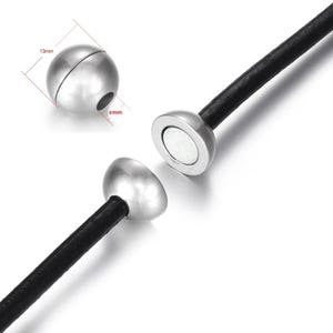 Pode incluir: Fecho magnético para joias, de cor prateada. O fecho tem um design esférico, com uma parte medindo 13 mm de diâmetro e 6 mm de profundidade. Está preso a um cordão preto.