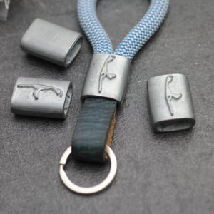 May include: A silver-toned keychain with a blue and white rope loop and a dark blue leather strap. The keychain features a metal accent with a stylized design. Additional metal pieces with the same design are also present.