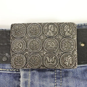 May include: A rectangular metal belt buckle featuring embedded coins. The buckle has a textured surface and is attached to a black leather belt. The coins display various designs, including portraits and text, with a vintage aesthetic.