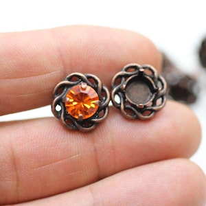 Pode incluir: Duas configurações de joias em forma de flor de cor de cobre. Uma configuração contém uma pedra preciosa redonda laranja. A outra configuração está vazia. As configurações são mantidas na mão de uma pessoa. As configurações são para fazer joias.