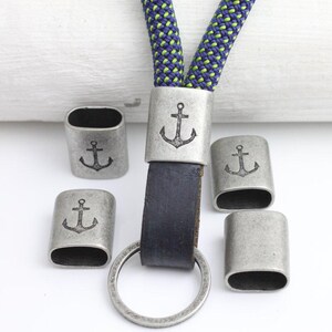 May include: A lanyard with a blue and green rope, a dark blue leather strap, and a silver-colored metal ring. The metal pieces have an anchor design. Several additional metal pieces with anchor designs are also present.