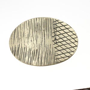 Peut inclure: Un objet décoratif ovale de couleur métallique. La surface est divisée en deux sections distinctes. Un côté présente des lignes verticales texturées, l'autre un motif quadrillé. L'objet a un aspect vintage ou antique.