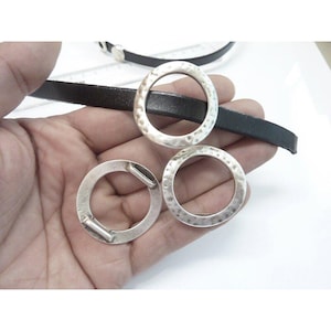Puede incluir: Tres anillos de metal plateado con textura martillada y una correa de cuero negro. Los anillos son circulares con una pequeña barra en un lado. La correa de cuero es fina y oscura. Los artículos se muestran sobre una superficie blanca.