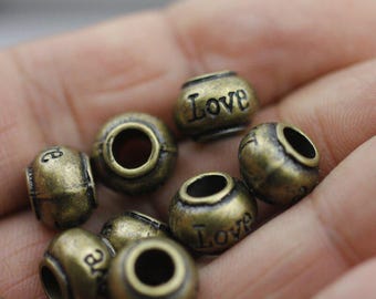 Może przedstawiać: Zbliżenie na kolekcję antycznych koralików w kolorze brązu. Niektóre koraliki mają wygrawerowane słowo "Love", inne litery. Koraliki są okrągłe z centralnym otworem, idealne do tworzenia biżuterii lub rękodzieła.