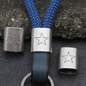 May include: Close-up of three silver-colored metal tube beads with a star design. The beads are on a blue and black rope lanyard with a dark blue leather strap and a silver keyring. The beads are cylindrical.