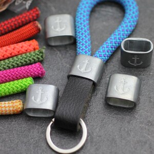 May include: A key fob with a black leather strap and a silver-colored metal anchor emblem. The key fob is attached to a blue and purple rope. Several colorful ropes are visible in the background, along with additional metal anchor emblems.