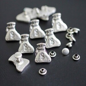 May include: A collection of silver-toned metal embellishments, including money bag charms, rivets, and a button-style fastener. The money bags feature a dollar sign. These small decorative elements are suitable for various craft projects.
