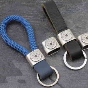 May include: Keychains with compass designs. One keychain features a blue rope loop and a blue leather strap. Another keychain has a black leather strap. Both have silver-colored metal accents with compass details and a silver key ring.