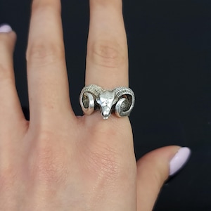 May include: A silver-toned ring featuring a detailed ram skull design with prominent curved horns. The metal has a textured, oxidised finish that highlights the contours of the skull and the spiralling ridges of the horns across the band.