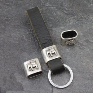 May include: A black leather keychain with a silver bulldog emblem and a silver keyring. Two additional silver bulldog emblems are also present. The keychain is designed for holding keys and is a dog-themed accessory.
