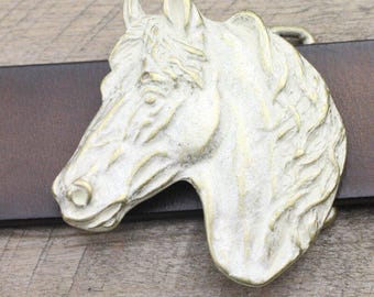 May include: A detailed, antique-style belt buckle featuring a horse head. The buckle is a light, aged gold color with intricate detailing, mounted on a brown leather belt. The horse head is sculpted with a weathered finish.