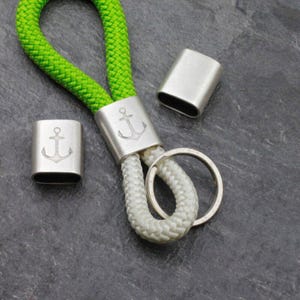 May include: A nautical-themed keychain featuring a green and white rope design. The keychain includes silver-colored metal accents with an anchor emblem. The design incorporates a key ring and two additional metal pieces.