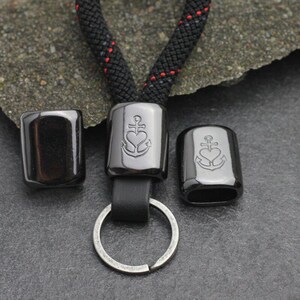 May include: A black lanyard with a key ring and three black metal beads. The center bead features an anchor inside a heart. The lanyard is made of black rope with red accents.