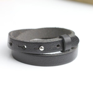 Könnte beinhalten: Ein schwarzes Wickelarmband aus Leder mit strukturierter Oberfläche, einem einzelnen silberfarbenen Metallniet, mehreren Einstelllöchern und einer passenden Lederschlaufe, um das Ende des Riemens in einer kreisförmigen Anordnung zu sichern.