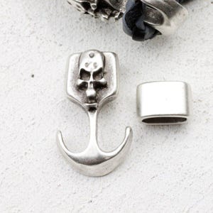 May include: Silver-toned metal anchor charm with a skull design, and a rectangular clasp. The anchor charm features a skull at the top and a curved anchor shape. The clasp is a simple, rectangular shape. The items are on a white surface.