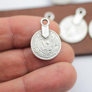 May include: A silver-colored coin charm with a loop for hanging. The coin features an embossed design with Arabic script and a central number. The charm is held in a hand, with other similar charms visible in the background.