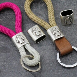 May include: Keychains with angel-themed metal accents. One keychain has a pink rope, another has a beige rope, and the third has a brown leather strap. The metal accents are silver-colored and feature an angel design. Each keychain has a silver-colored key ring.