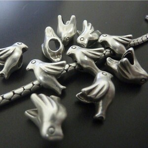 Op de afbeelding: Een verzameling zilverkleurige vogelvormige kralen, sommige met open gaten, ligt op een donkere ondergrond. De kralen zijn gedetailleerd met vleugels en kleine oogmarkeringen. Een zilveren ketting is ook zichtbaar, met de vogelkralen eraan bevestigd.